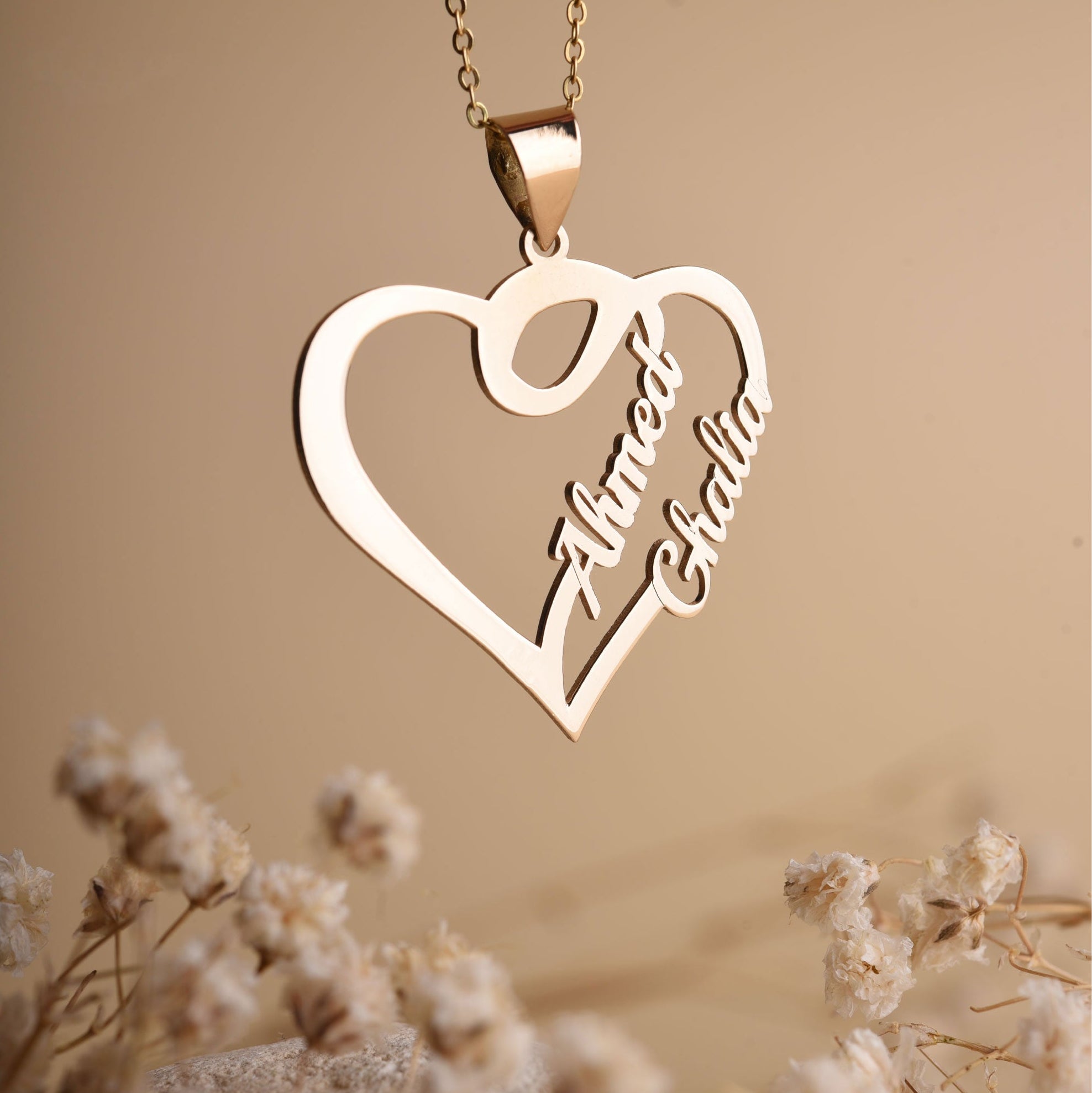 Gold heart-shaped necklace with engraved names on a beige background with dried flowers.