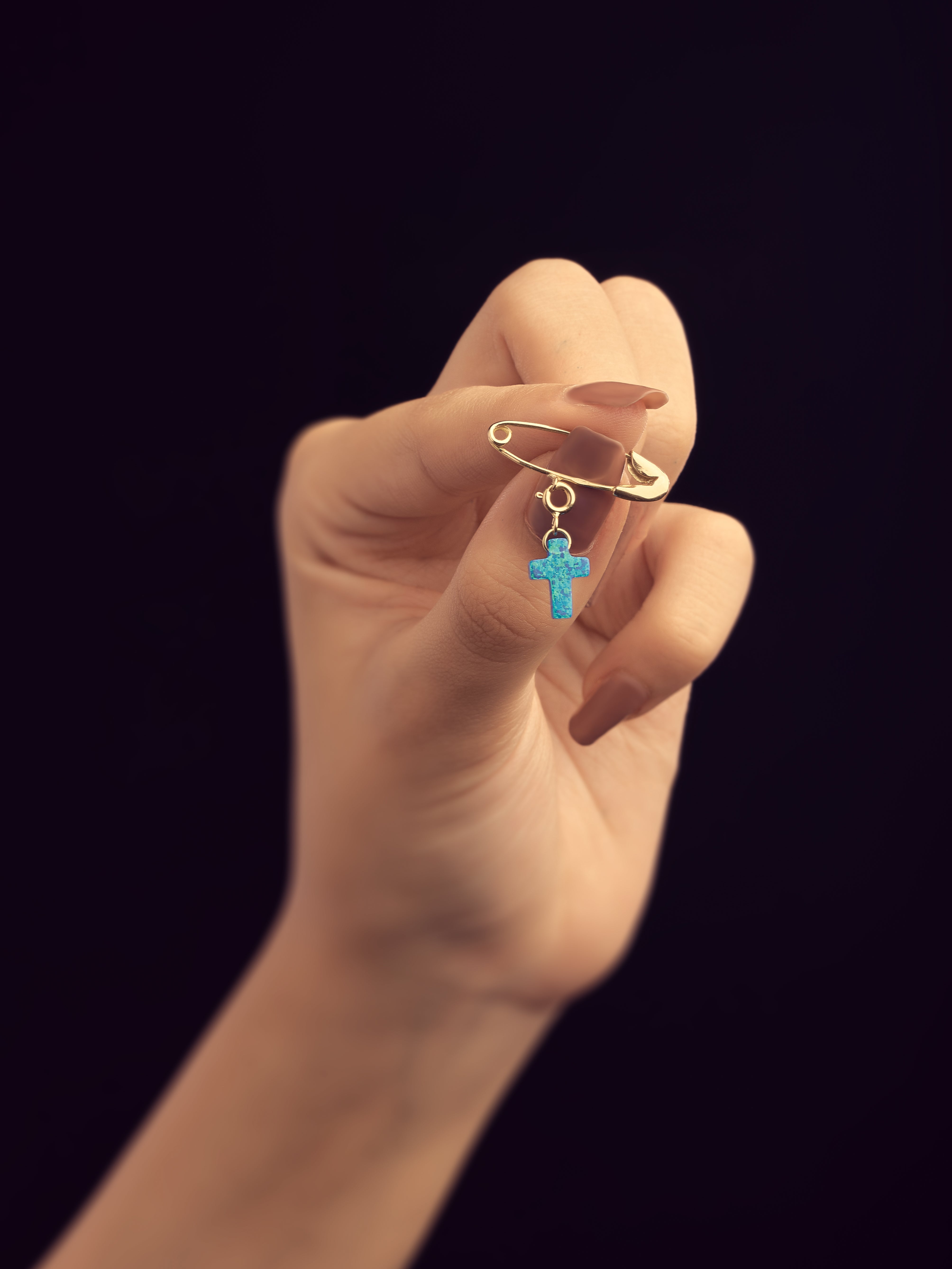 Hand holding a safety pin with a blue cross pendant against a black background